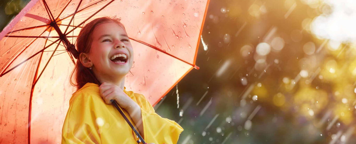 schulmaedchen-lacht-vor-vergnuegen-unter-lichtdurchflutetem-roten-regenschirm-in-gelber-regenjacke-tropfen-reflektieren-das-sonnenlicht